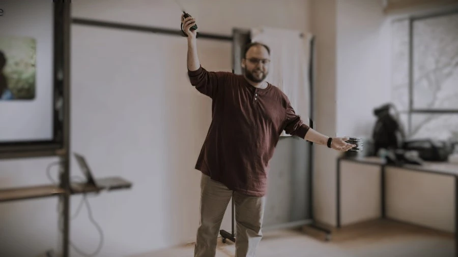 A male speaker enthusiastically gesturing with his right arm raised and holding a remote or pointer, while his left arm is extended to the side. He is smiling and leaning slightly forward, demonstrating confident, open, and dynamic body language during a presentation