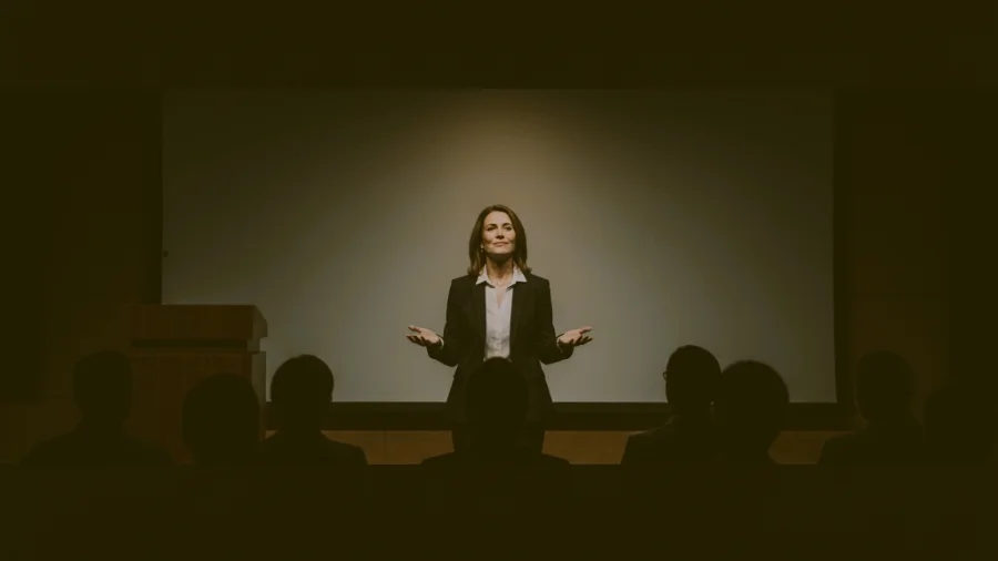 A confident female speaker standing still on a stage with her arms open and palms up in a relaxed, controlled gesture, utilizing a deliberate pause to command the quiet attention of the silhouetted audience.